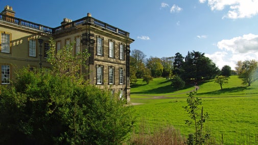 A view of the west side of the house and a glimpse of the Pleasure Grounds through trees on a sunny day at Calke Abbey